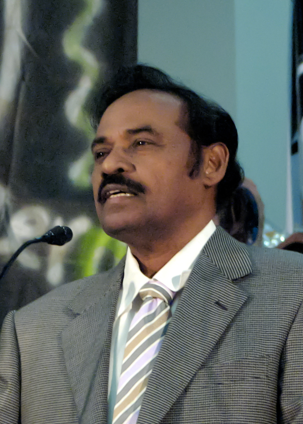 Dr. T. R. Paarivendhar speaking into a microphone, wearing a grey checked jacket and striped tie