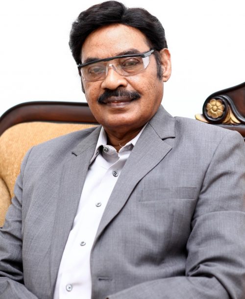 Portrait of Dr. T. R. Paarivendhar in a grey suit and open-collared shirt, seated on a carved wooden sofa