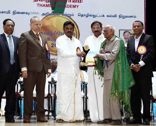 Dr. T. R. Paarivendhar with dignitaries on stage as a trophy is presented to an elderly honoree wearing a green shawl, beneath a Thamizh Academy banner