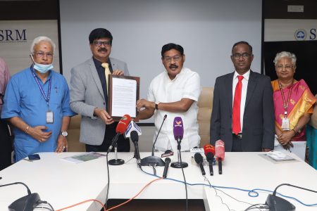 Dr. T. R. Paarivendhar, in a white shirt, holding a signed document with a colleague at a table of media microphones in an SRM hall