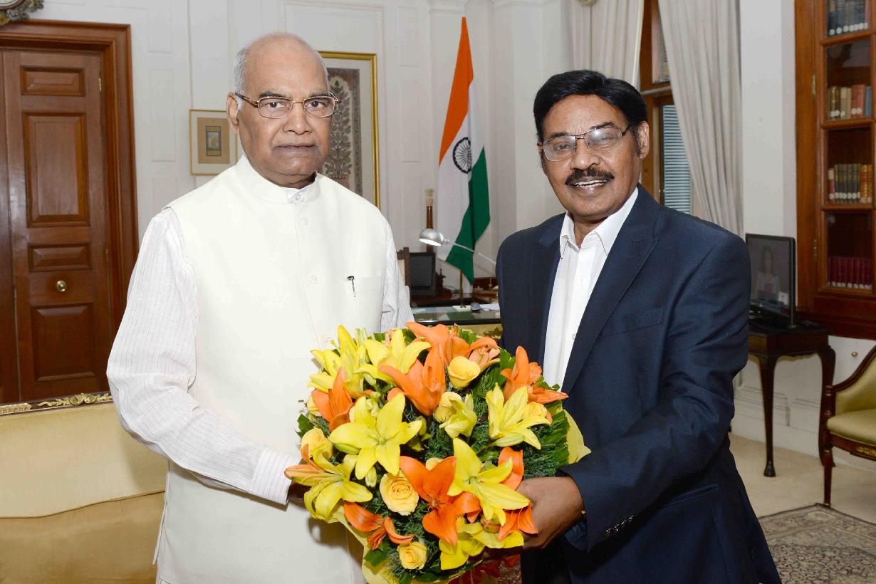 Dr. T. R. Paarivendhar in a dark suit presenting a bouquet of yellow and orange lilies to a senior dignitary in white, in a formal office with the Indian national flag