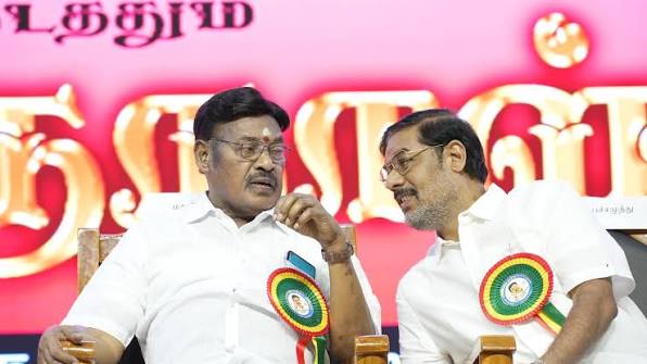 Dr. T. R. Paarivendhar seated on a stage in a white shirt and rosette badge, speaking with another man, in front of a pink and gold banner with Tamil lettering