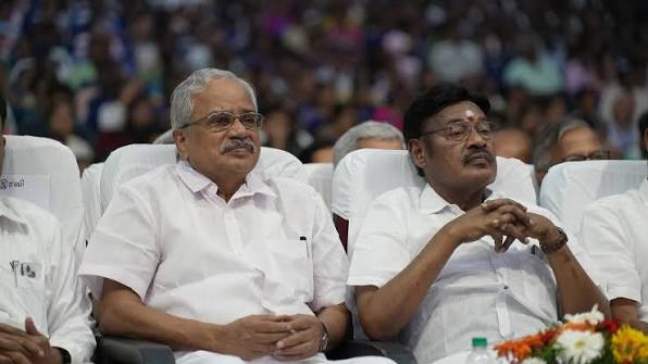 Dr. T. R. Paarivendhar seated in the front row of a large public programme beside another guest, both in white shirts, with a crowded audience behind