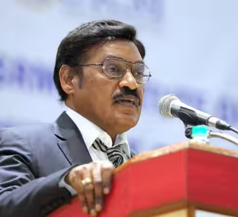 Dr. T. R. Paarivendhar in glasses and a dark suit speaking at a red lectern with a microphone